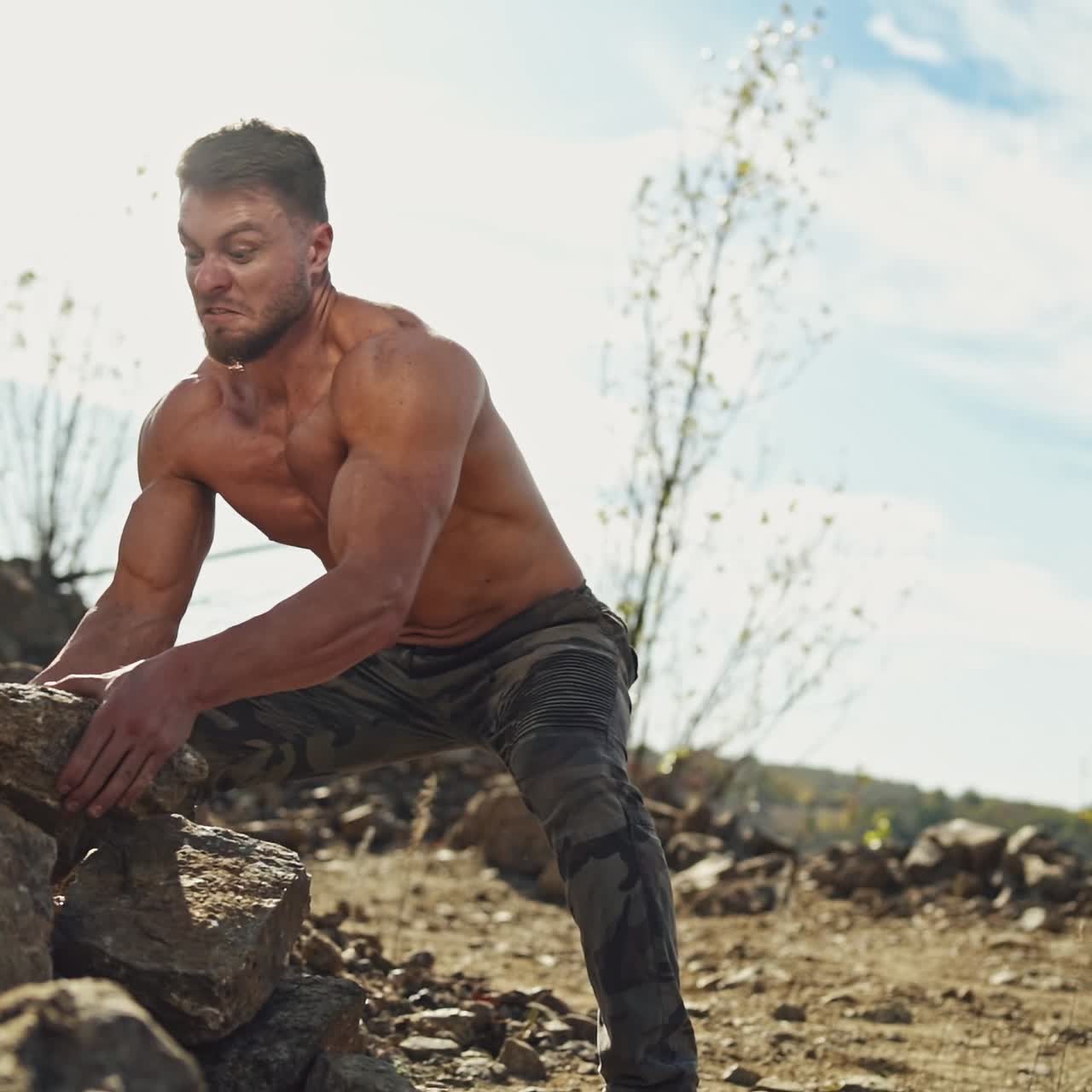 Angry sportsman with muscular body throws stones in nature. Shirtless bodybuilder exercises with heavy stones outdoors. Slow motion.