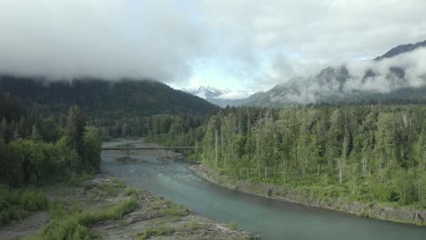 Drone-footage-of-beautiful-Pacific-Northwest-river,-misty-forest-with-bridge-and-mountains-in-background