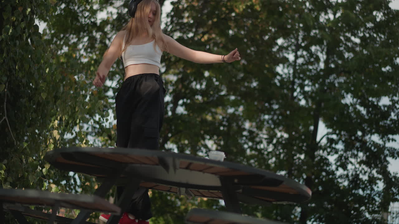 Donna bianca che balla su un tavolo da picnic, gioiosa performance urbana con cuffie e luci decorative al crepuscolo, alberi e sfondo di edifici cittadini, abbigliamento casual con canottiera e pantaloni ampi, movimento espressivo