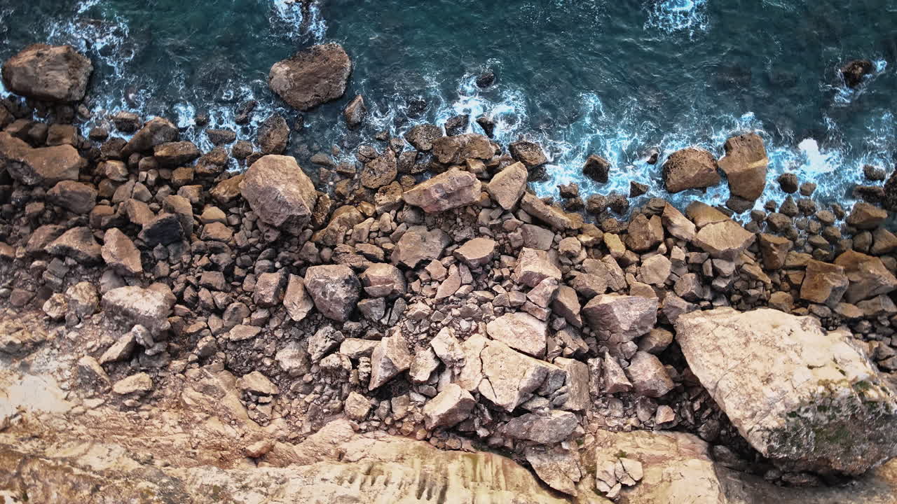 La telecamera esegue una panoramica in senso orario su una costa rocciosa spettacolare dove le onde si infrangono sui massi, catturando l'energia selvaggia dell'Atlantico di Sagres