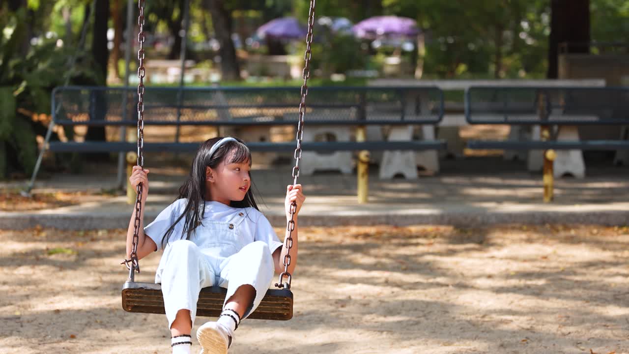 Una giovane ragazza asiatica che indossa una salopette di jeans si diverte a dondolarsi in un parco giochi all'aperto e soleggiato.