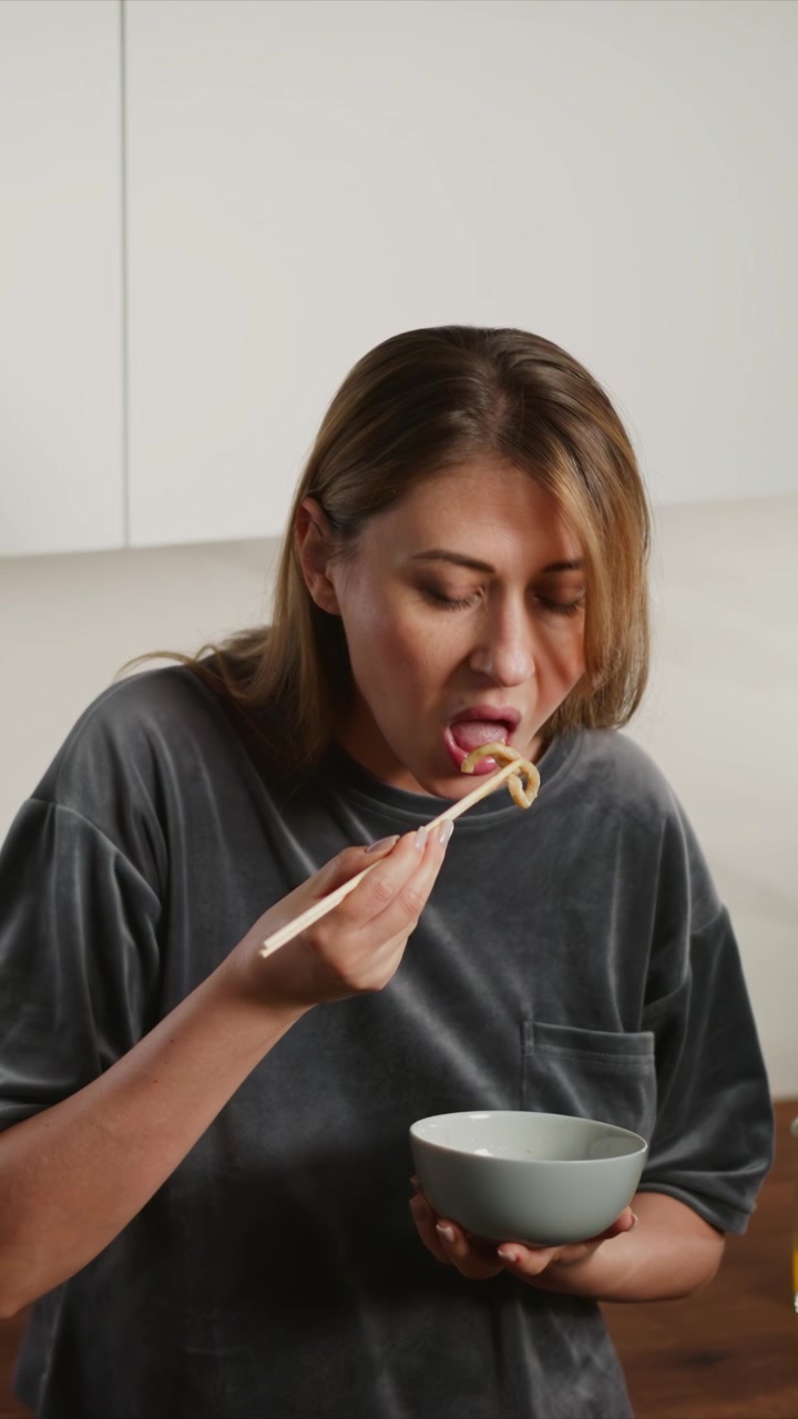 Close-up de mulher comendo macarrão com pauzinhos enquanto segura uma tigela nas mãos. A cena apresenta iluminação suave e movimento mínimo da câmera, focando na ação contra um fundo simples. Vidro