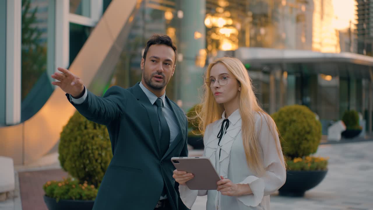 Businessman Leader Giving Directions to Young Woman Trainee Looking at Digital Tablet Outside Office Building at Sunset, slow motion, medium shot