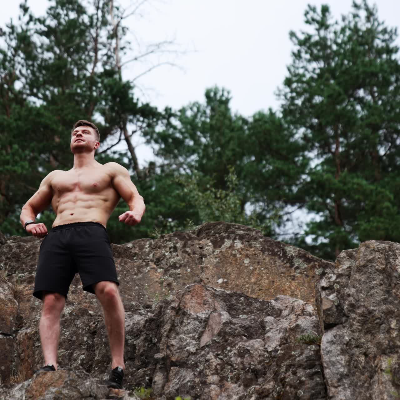 Athletic man with naked torso wearing black shorts stands on the rock. Sportsman rises hands showing muscles. Low angle view