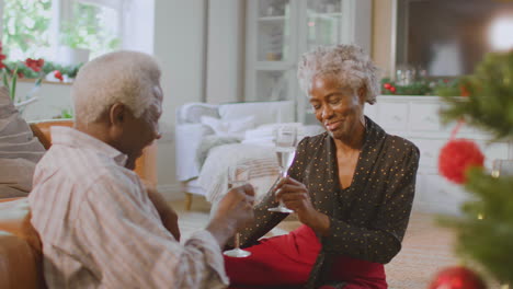 Amorosa-Pareja-De-Ancianos-Celebrando-Con-Champán-Alrededor-Del-árbol-De-Navidad-En-Casa