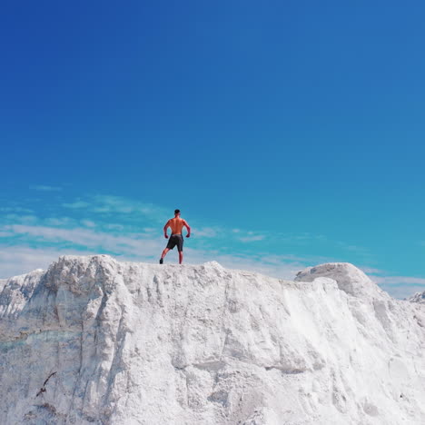 drone view on the high white mountain and muscular man on top of it in summer. topless young sportsman showing his trained body while standing on the top of the hill under blue sky.