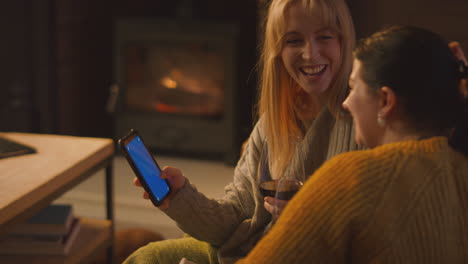 Dos-Amigas-Relajándose-En-El-Sofá-Mirando-El-Teléfono-Móvil-Con-Un-Acogedor-Fuego-Sosteniendo-Una-Copa-De-Vino