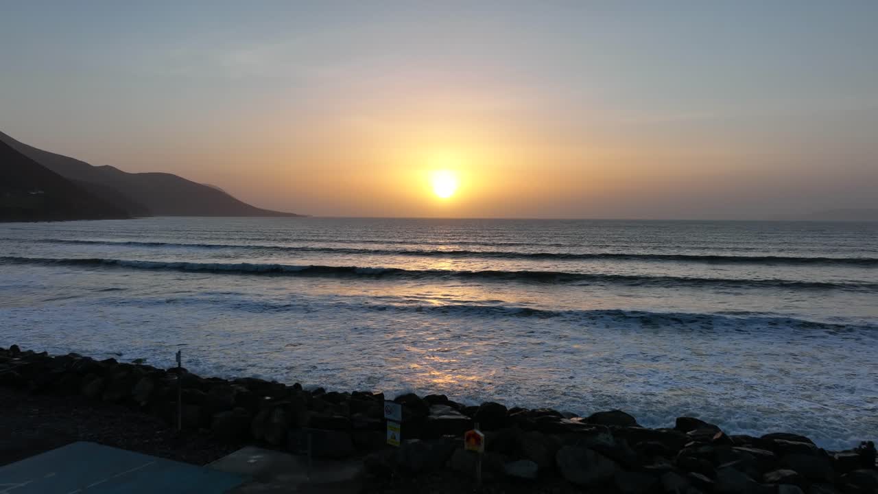 Rossbeigh Beach, County Kerry, Ireland, March 2022. Drone pushes west out over the car park and rocks passing above smooth rolling ocean waves as the sunset light shimmers on the water.