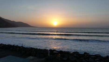 Rossbeigh Beach, County Kerry, Ireland, March 2022. Drone pushes west out over the car park and rocks passing above smooth rolling ocean waves as the sunset light shimmers on the water.