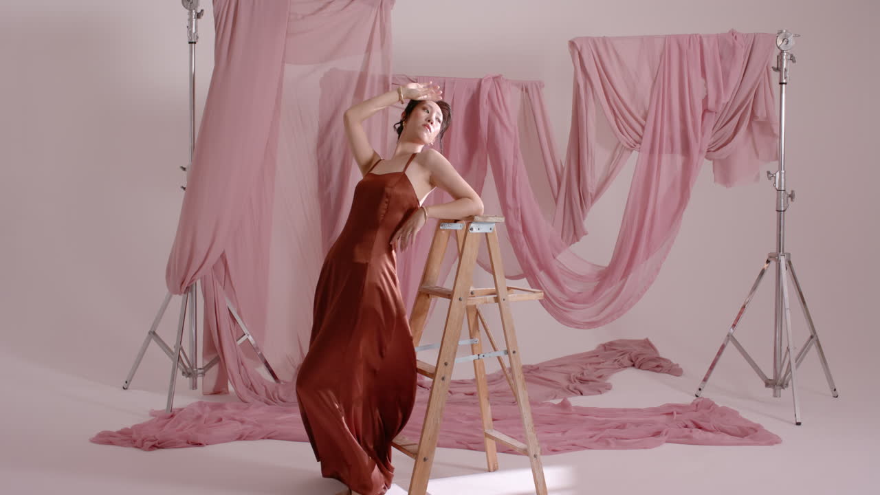 Asian woman in satin burgundy dress leaning pivoting on wooden stepladder in studio fashion shoot