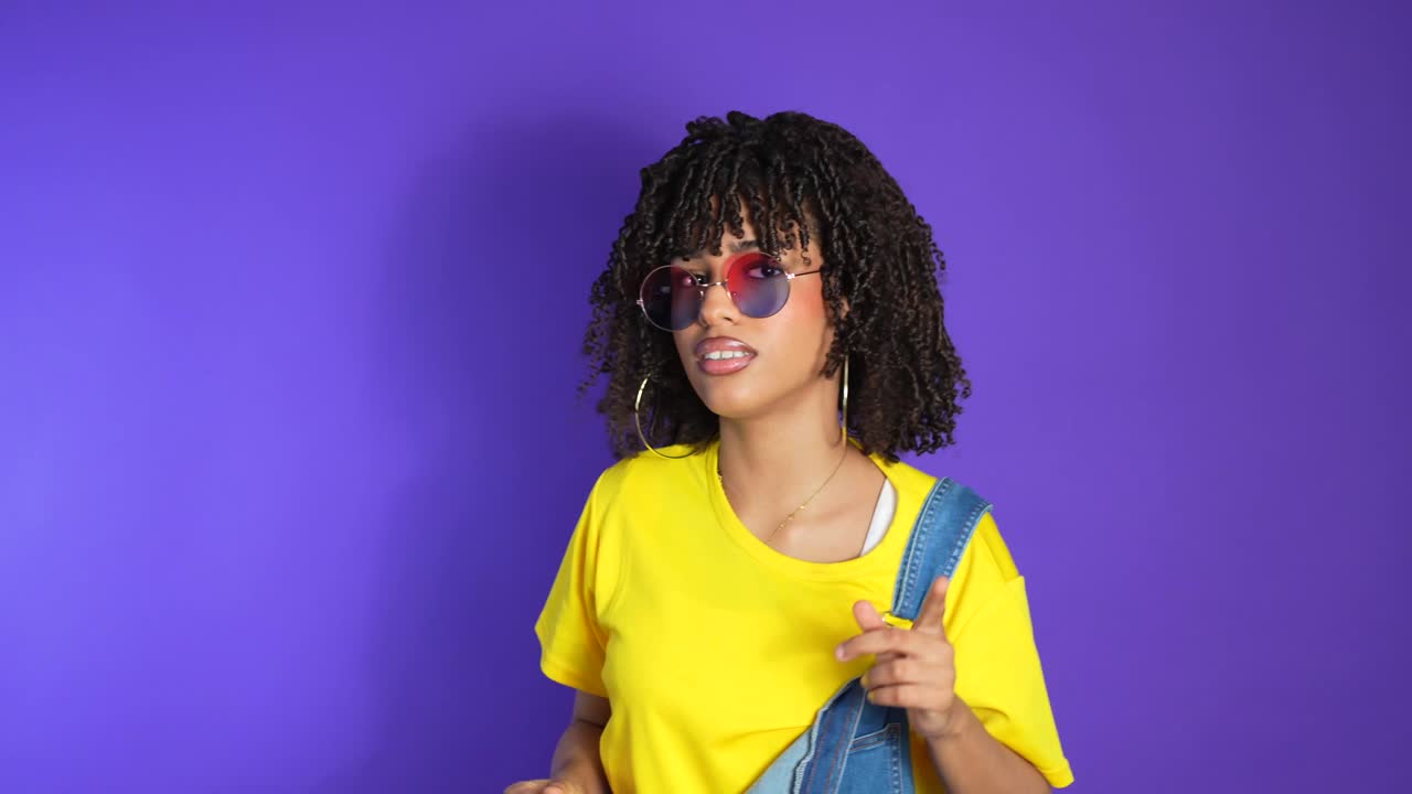 A woman in a yellow t-shirt and overalls posing in front of a purple background