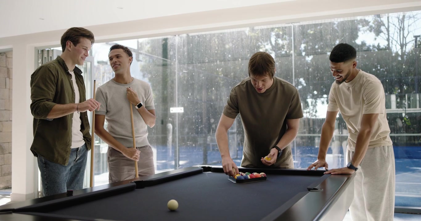 Diverse men gathering at black felt pool table, centered man placing rack, arranging balls for play