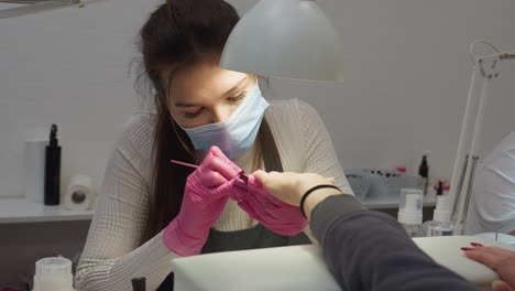 nail technician wearing mask focused on carefully applying polish to client nail under lamp in modern salon while another person arm appears in foreground, highlighting attention to detail