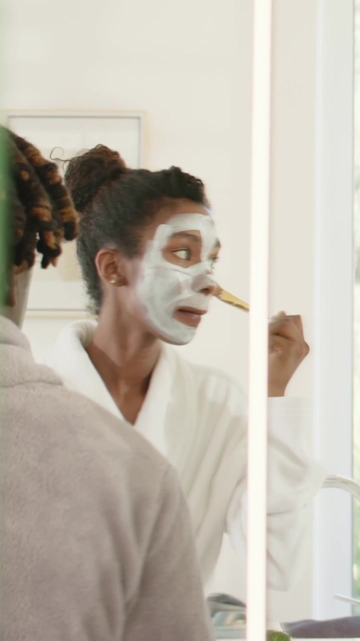 Video vertical: Mujer con bata blanca aplicándose una mascarilla con brocha en el lavabo, mientras se mira al espejo para revisar su piel.