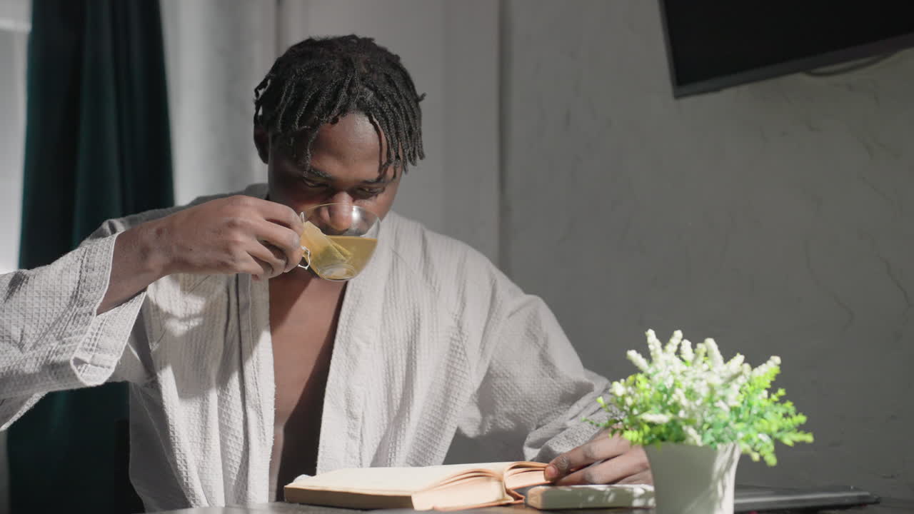 Man in bathrobe sitting at table, reading book and drinking tea. Indoor lifestyle scene with peaceful morning ambiance. Soft lighting, plant on table adds to calm, cozy atmosphere