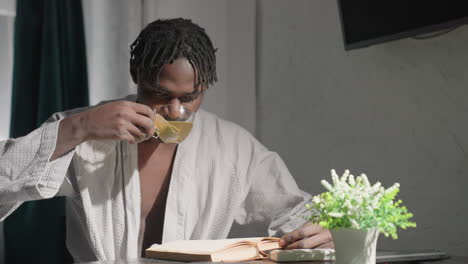 man in bathrobe sitting at table, reading book and drinking tea. indoor lifestyle scene with peaceful morning ambiance. soft lighting, plant on table adds to calm, cozy atmosphere