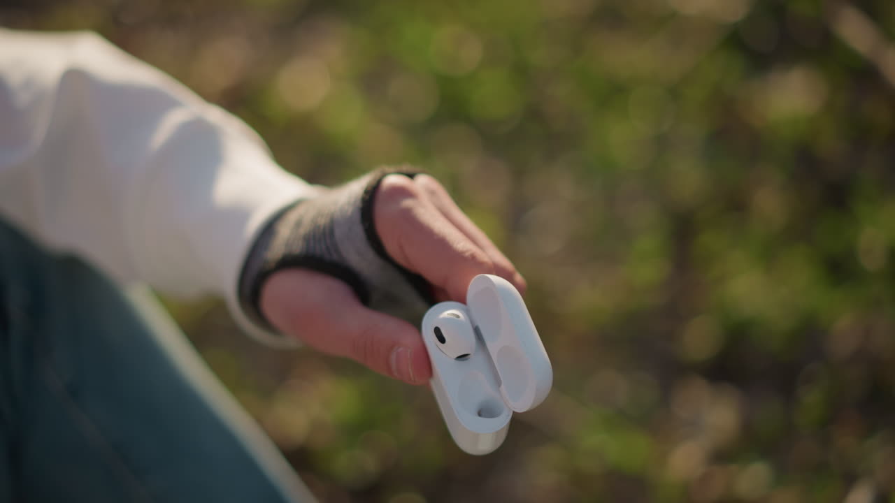 이어폰을 낀 여자가 풀밭에서 케이스에서 이어폰을 꺼내는 모습이 오후 햇살에 비치고 있어요. 편안해 보이는 여자가 부드러운 햇살 아래 푸르른 녹음 속에서 케이스에서 이어폰을 살짝 꺼내고 있습니다.