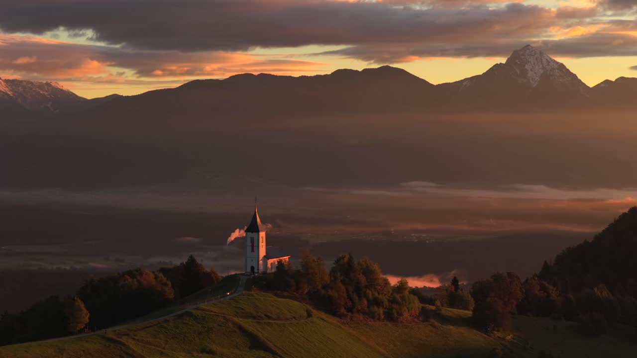 Aerial sunrise view of Jamnik church on a hill, serene with Alps backdrop