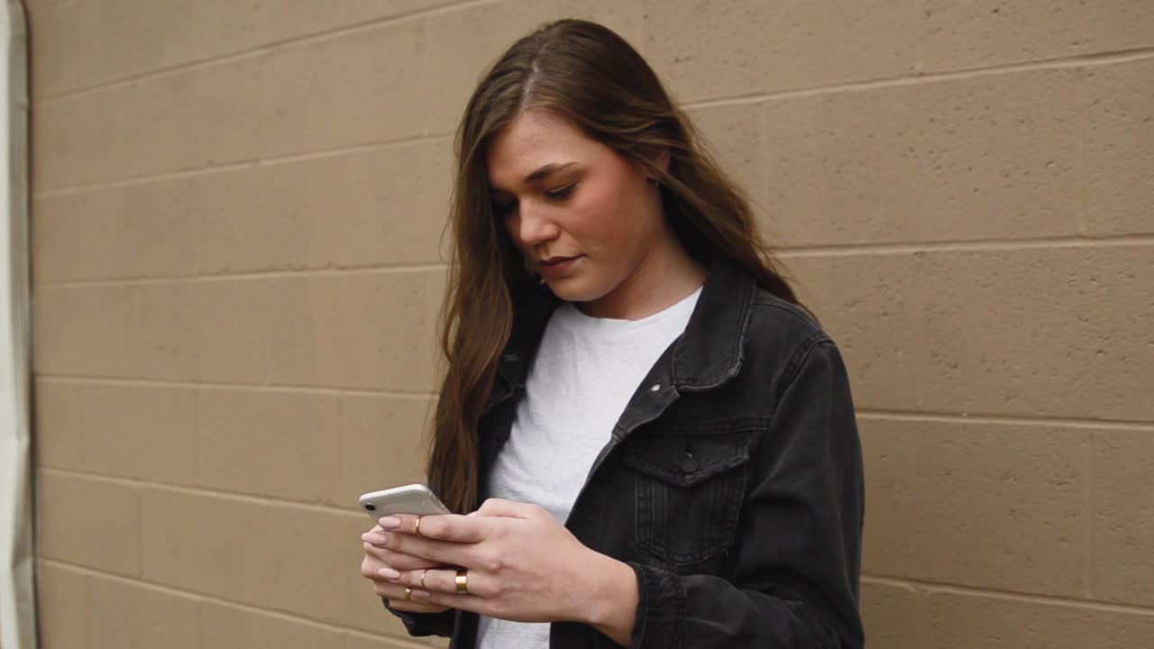 Pull away steadicam shot of a beautiful,young, brunette college teenager texting on a cell phone wearing a white shirt with a black jacket against a wall