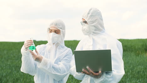 caucasian researchers in protective suit holding a test tube and using laptop while doing pest control in the green field