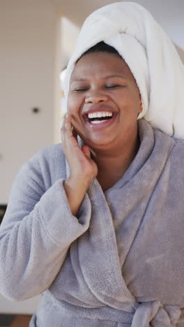 vertical video of african american woman in bathrobe with towel on head laughing in bathroom