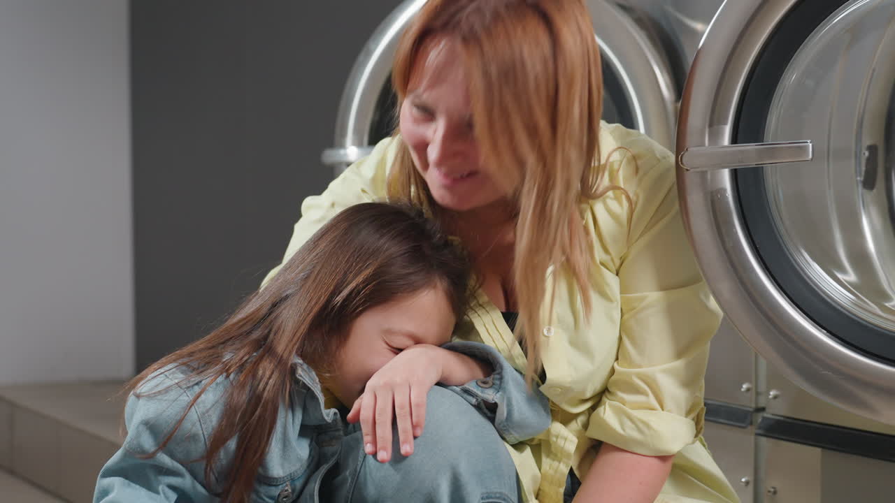Business woman with daughter rest and play beside industrial washers inside laundromat after cleaning, smiles on faces, open stainless door visible, warm family bonding moment