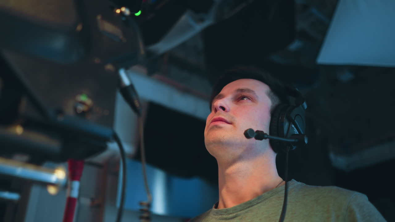 Cameraman wearing headset looks upward in concentration while operating television broadcast camera in studio environment, surrounded by production cables, equipment, and soft lighting during active filming session