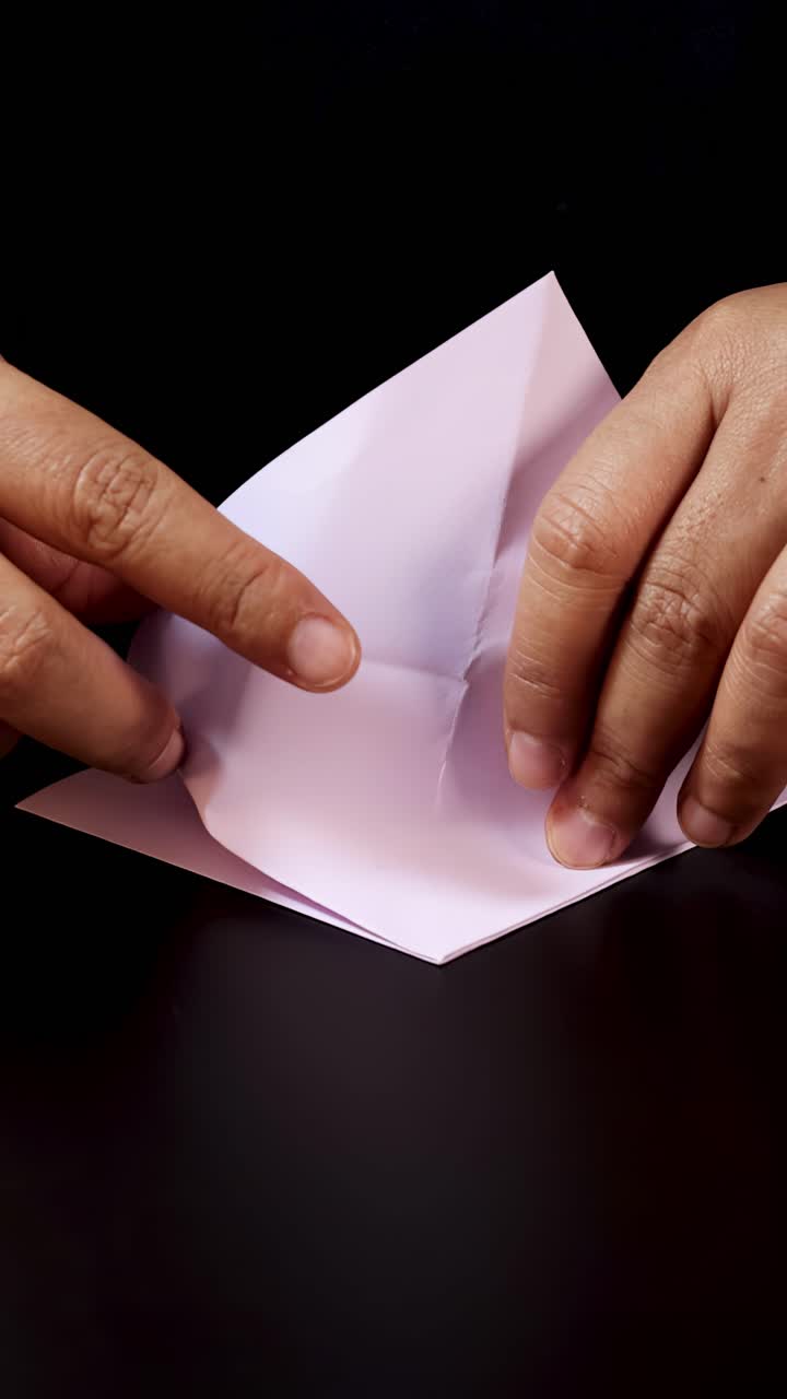 Mani adulte piegano con attenzione carta pastello in origami, in primo piano, sotto una morbida illuminazione da studio.