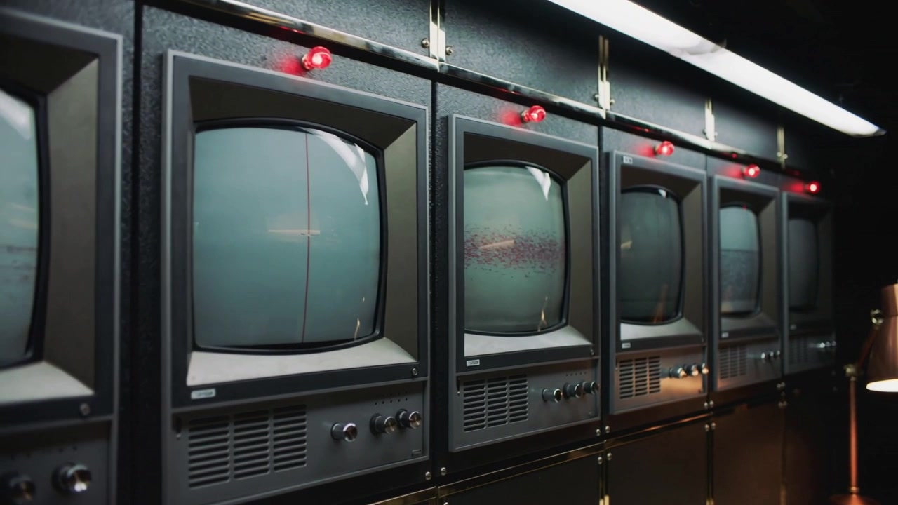 Panorerande kamera rör sig längs en rad vintage CRT-skärmar i ett dunkelt kontrollrum, som avslöjar röda lampor.