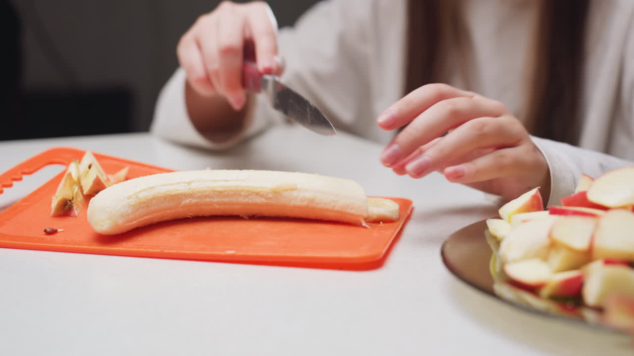 Hand view of girl cutting banana with knife on orange rubber board, peeled banana resting, apple scraps nearby, plate with apple slices on countertop, indoor light, shallow depth of field