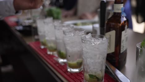 Bartender-pouring-drinks-into-a-row-of-iced-glasses-with-mint-and-lime-at-a-bar