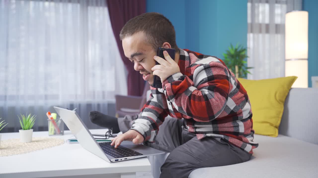 Happy and cheerful dwarf young man working from home at laptop and talking on phone.
