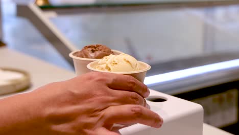 a hand reaches for and lifts a paper cup of ice cream from a modern display stand in a brightly lit dessert shop, with smooth camera movement