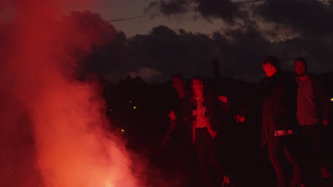 group of teenagers hanging out and dancing in light of red signal flare outdoors.