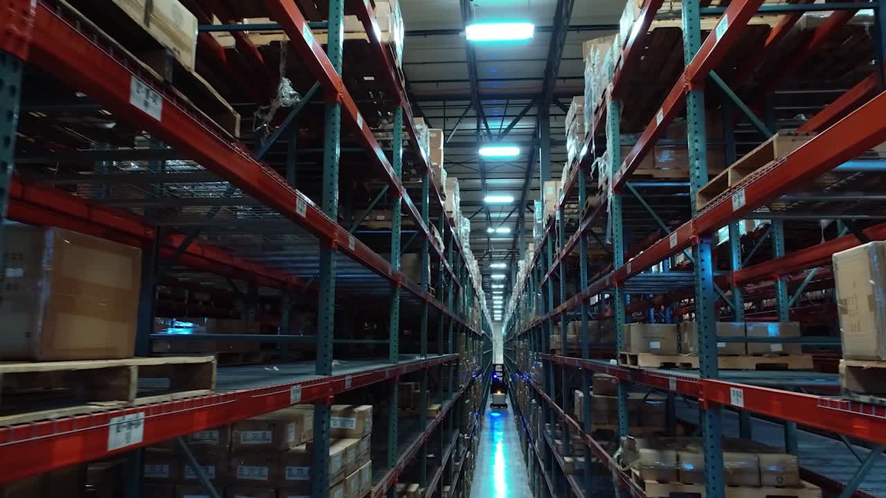 A cinematic drone shot flying down a tall warehouse aisle, smoothly tracking a forklift moving between shelves of stored packages