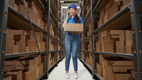 full body of asian female courier in blue uniform talking on smartphone while delivering a carton in warehouse