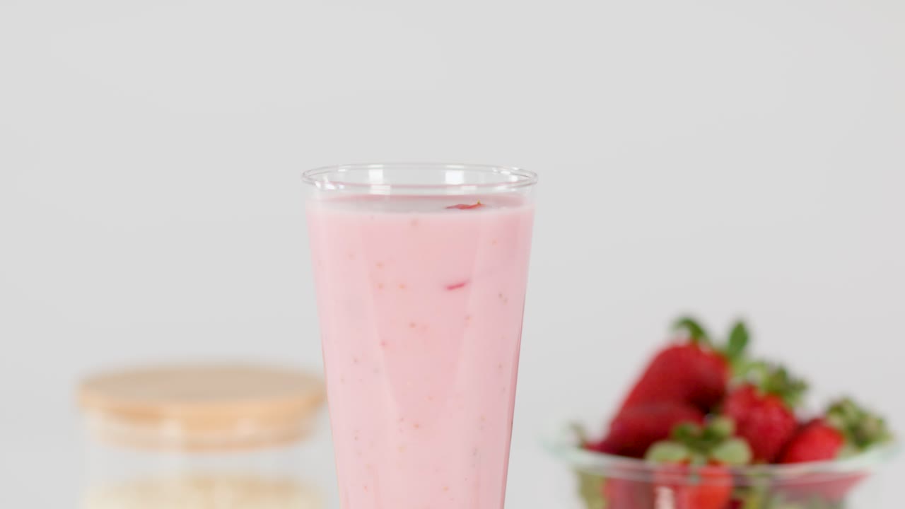 Hand places striped straw in creamy strawberry milkshake, bright background, fresh strawberries, static camera
