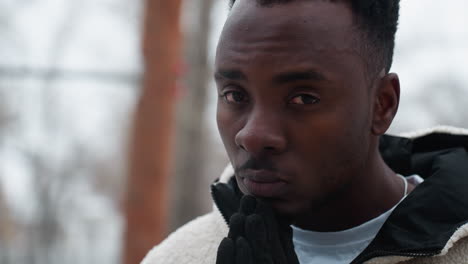 close-up of a reflective young boy wearing black gloves and white winter jacket, standing outdoors during winter with blurred background of bare trees and chilly weather