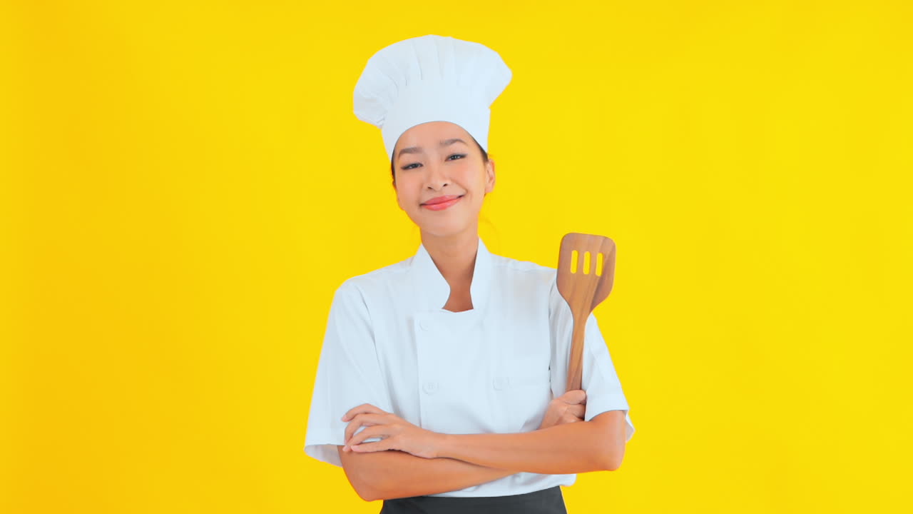 Seorang Koki Wanita Menarik dalam Seragam Koki Tradisional Berdiri dengan Latar Belakang Polos, Lengan Terlipat, dengan Spatula Kayu di Tangan