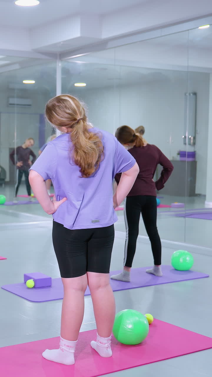 instructor de gimnasia conduce a tres mujeres a través de un ejercicio de torsión del cuello con las manos en la cintura, los participantes se paran en esteras de colores, rodeados de bolas de ejercicio y bloques, con espejos que reflejan la sesión