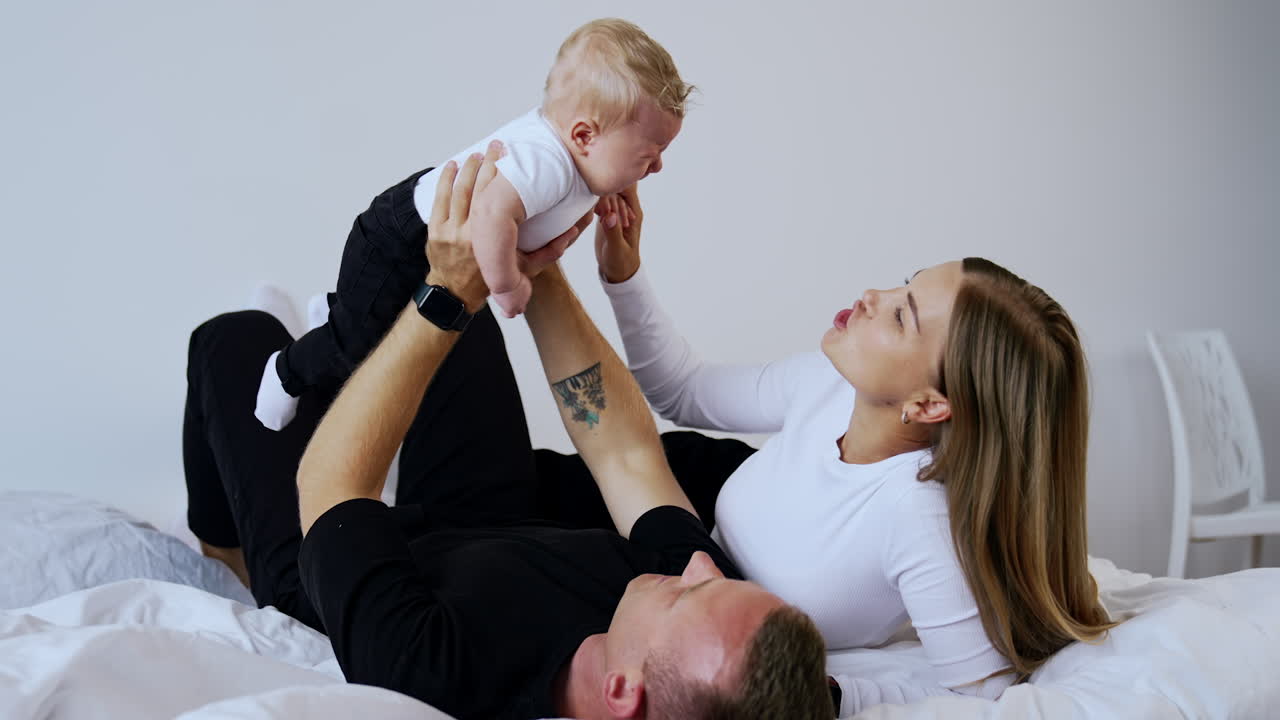 Father wearing black clothes lies on the bed holding a baby above him. Infant starts to cry and mother soothes him.