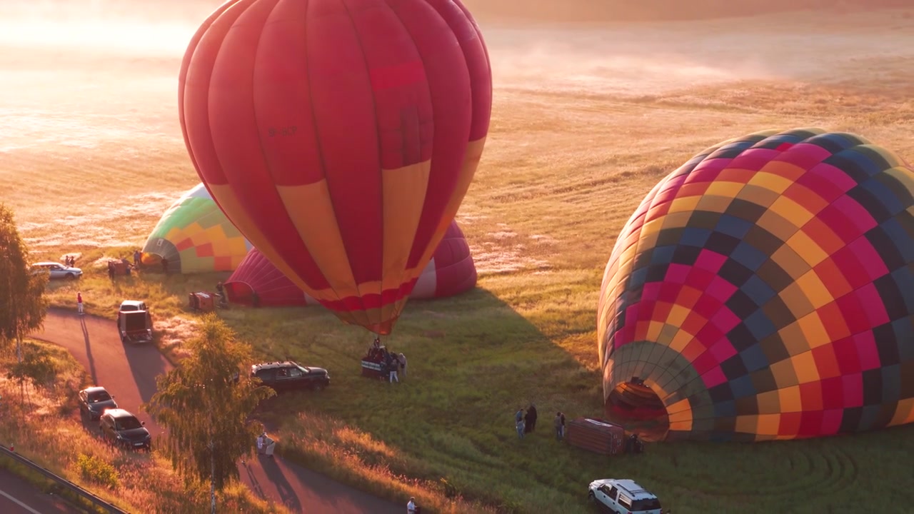 Toma amplia cinematográfica de globos aerostáticos coloridos inflándose en un campo de hierba durante un amanecer brumoso, capturando la magia del vuelo matutino y los viajes de aventura.