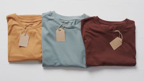 video of flat lay of multi coloured t shirts with tags and copy space on white background