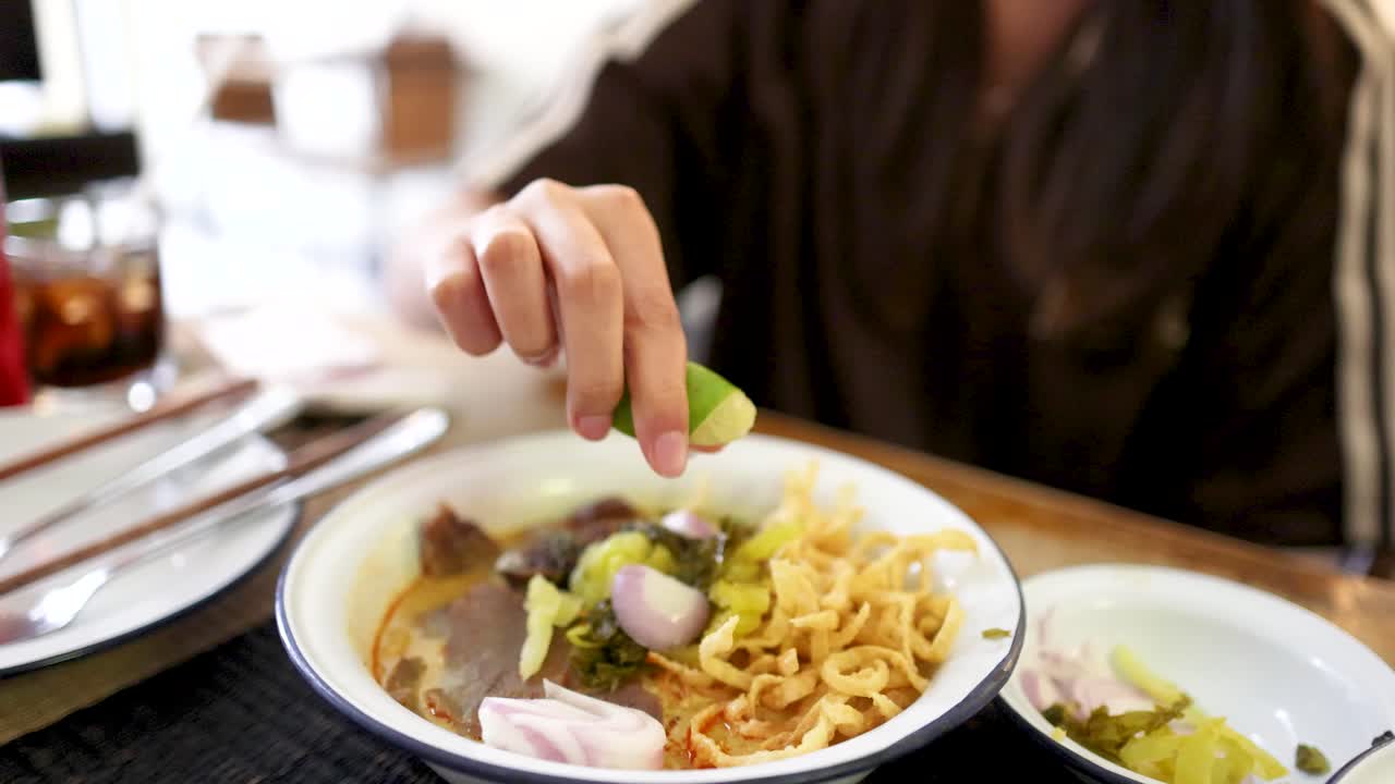 A person squeezes fresh lime onto a bowl of Northern Thai beef curry noodles in a well-lit Bangkok restaurant, captured in a close-up perspective