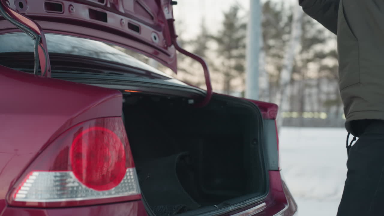 young man in winter jacket arranges items in boot of parked car then closes boot and walks away down snow-dusted road with blurred tree line and residential background