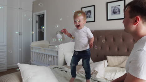 Caring-father-plays-son-looking-at-soap-bubbles,-the-boy-is-happy-jumping-on-the-bed