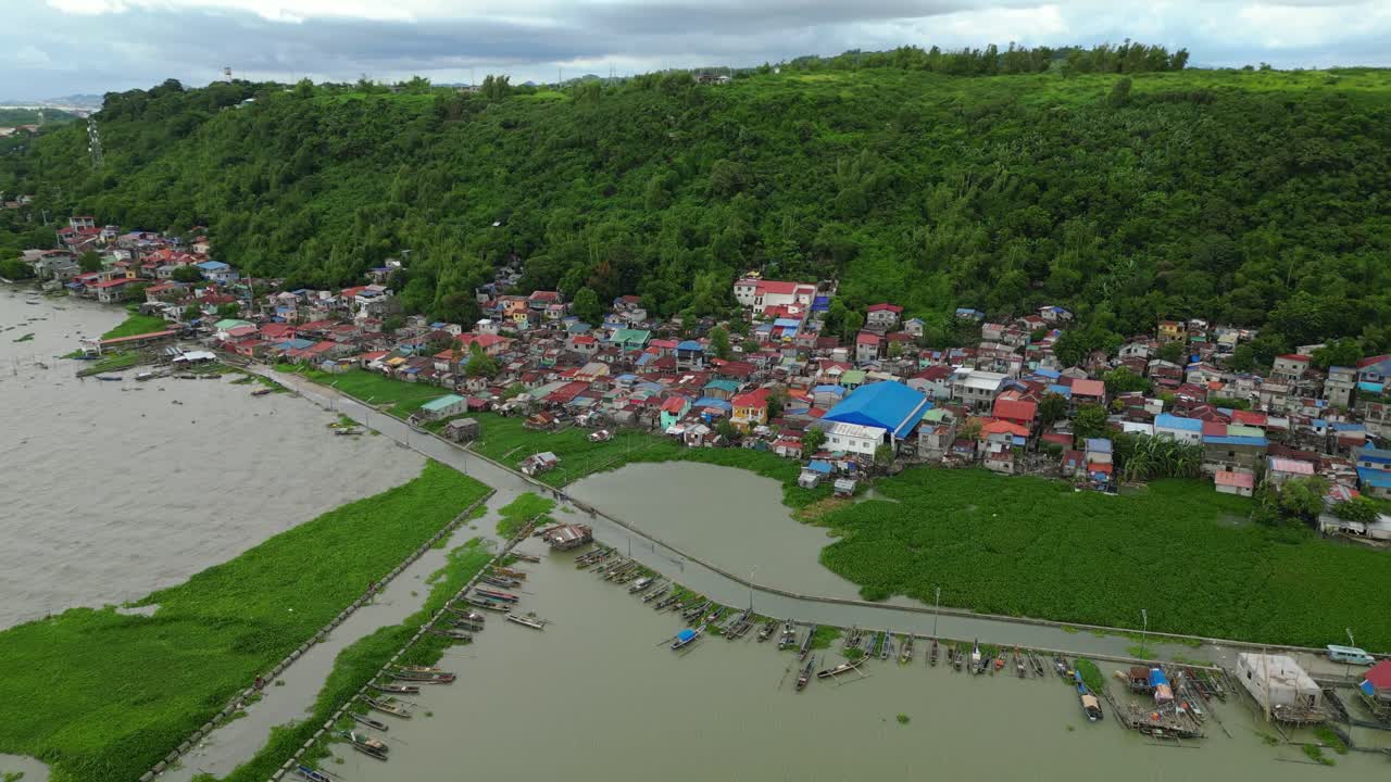 Drone shot captures a Philippine riverside village with vibrant rooftops, docked boats, and a forested hill backdrop, highlighting harmony between nature and settlement