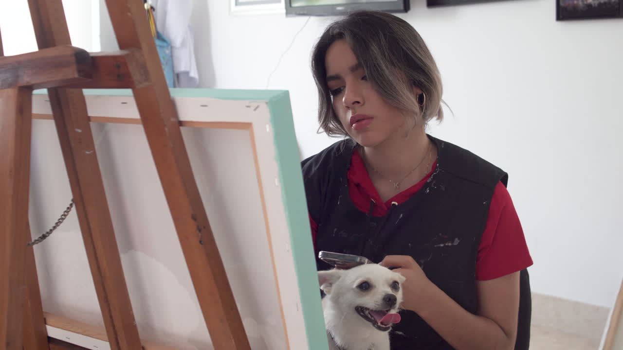 Attractive woman painting on canvas and looking for ideas in smartphone. Dog on her laps. Reveal shot