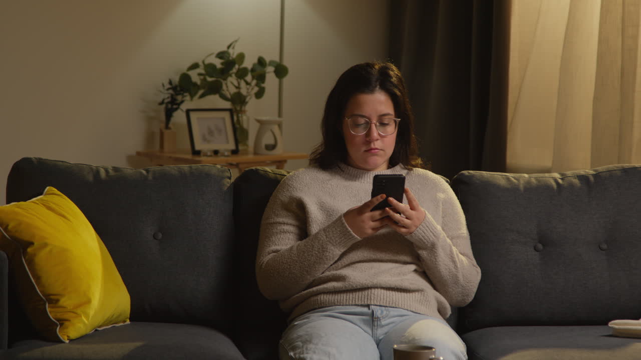 Young Woman Spending Evening At Home Sitting On Sofa With Mobile Phone Scrolling Through Internet Or Social Media