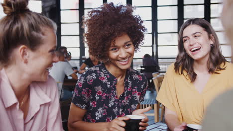 Cuatro-Jóvenes-Amigas-Reunidas-Sentadas-A-La-Mesa-En-Una-Cafetería-Y-Hablando